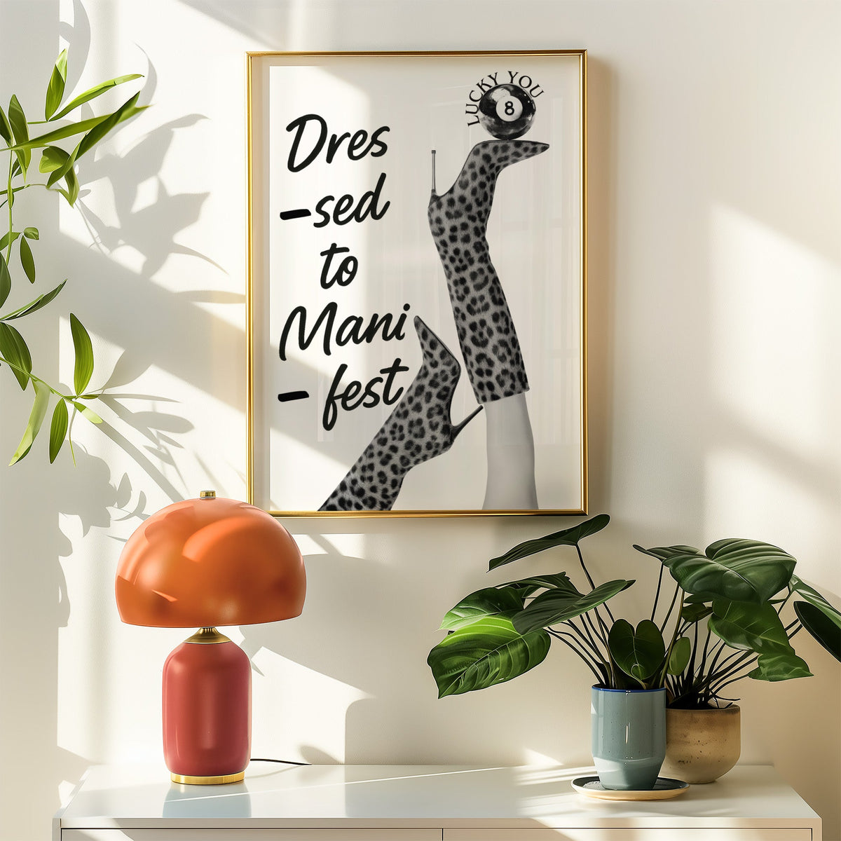 Legs in leopard boots balancing eight ball quote print shown in a gold frame above a white sideboard with an orange lamp