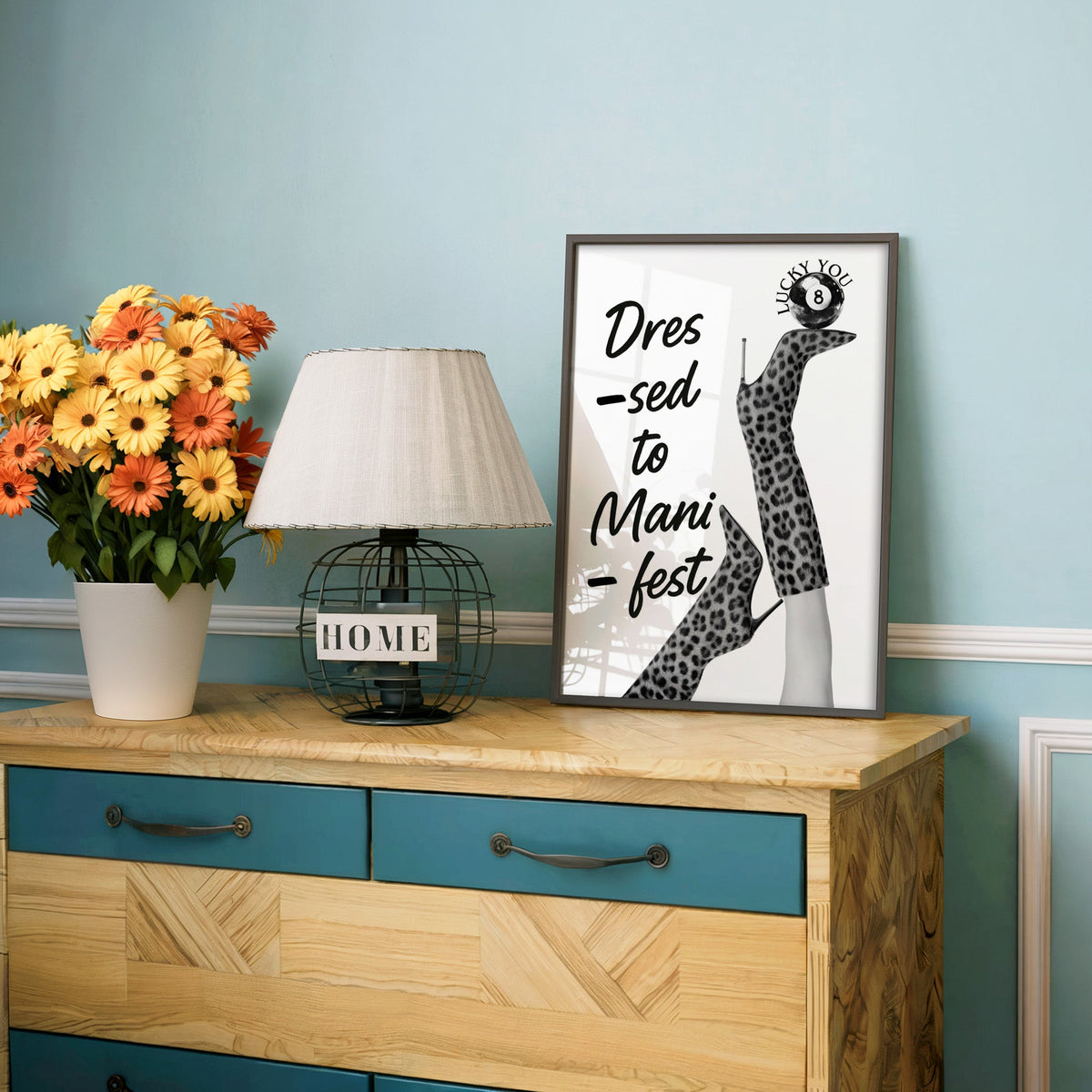 Legs in leopard boots balancing eight ball quote print in a charcoal frame on a teal dresser beside orange daisies