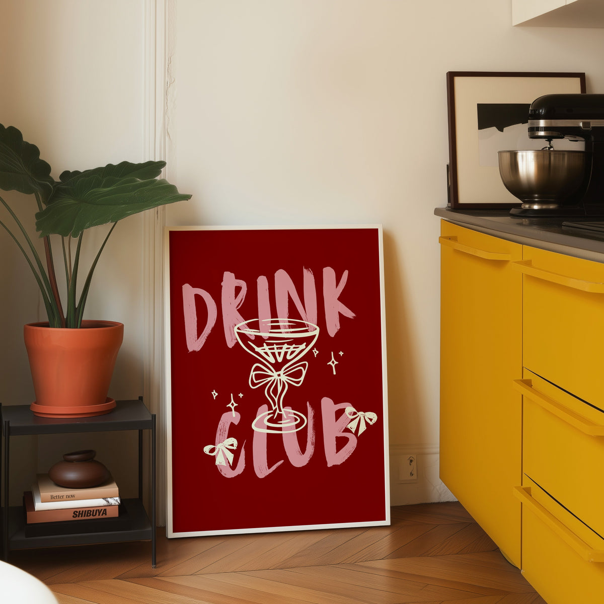 Drink Club print in a white frame leaning on parquet beside a bright yellow kitchen
