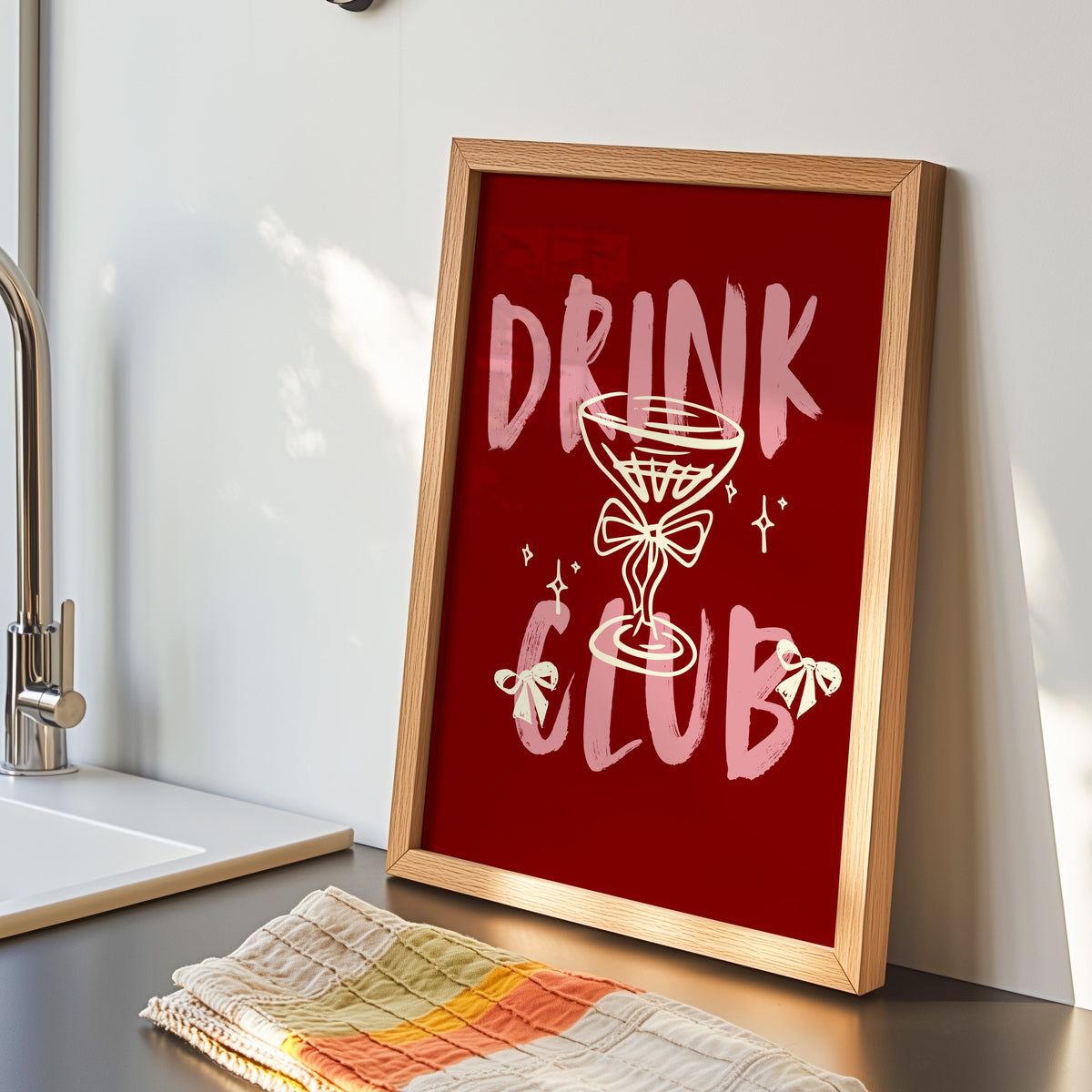 Drink Club print in an oak frame by the kitchen sink beside a striped tea towel