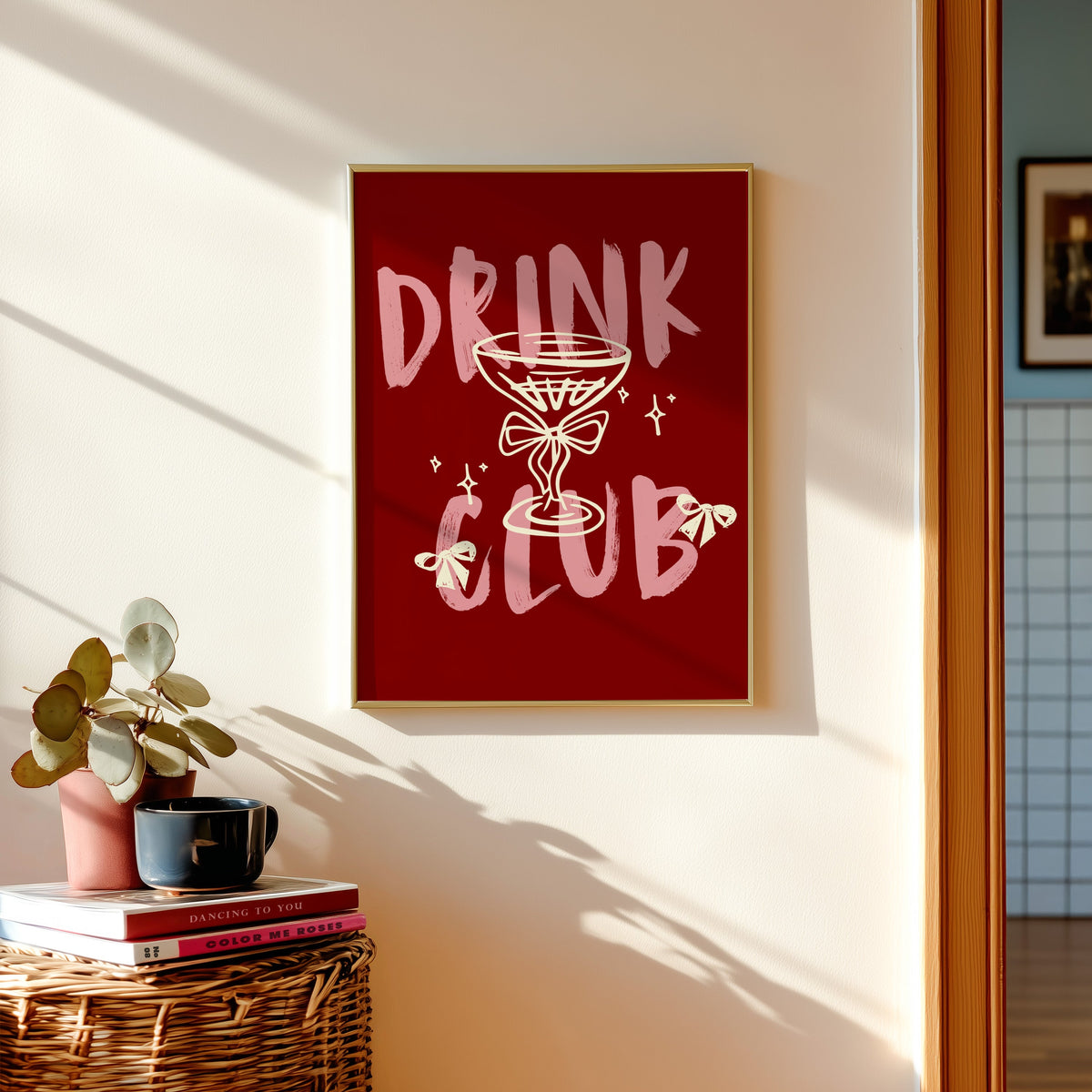 Drink Club print shown in a gold frame above a basket of books and a pilea plant