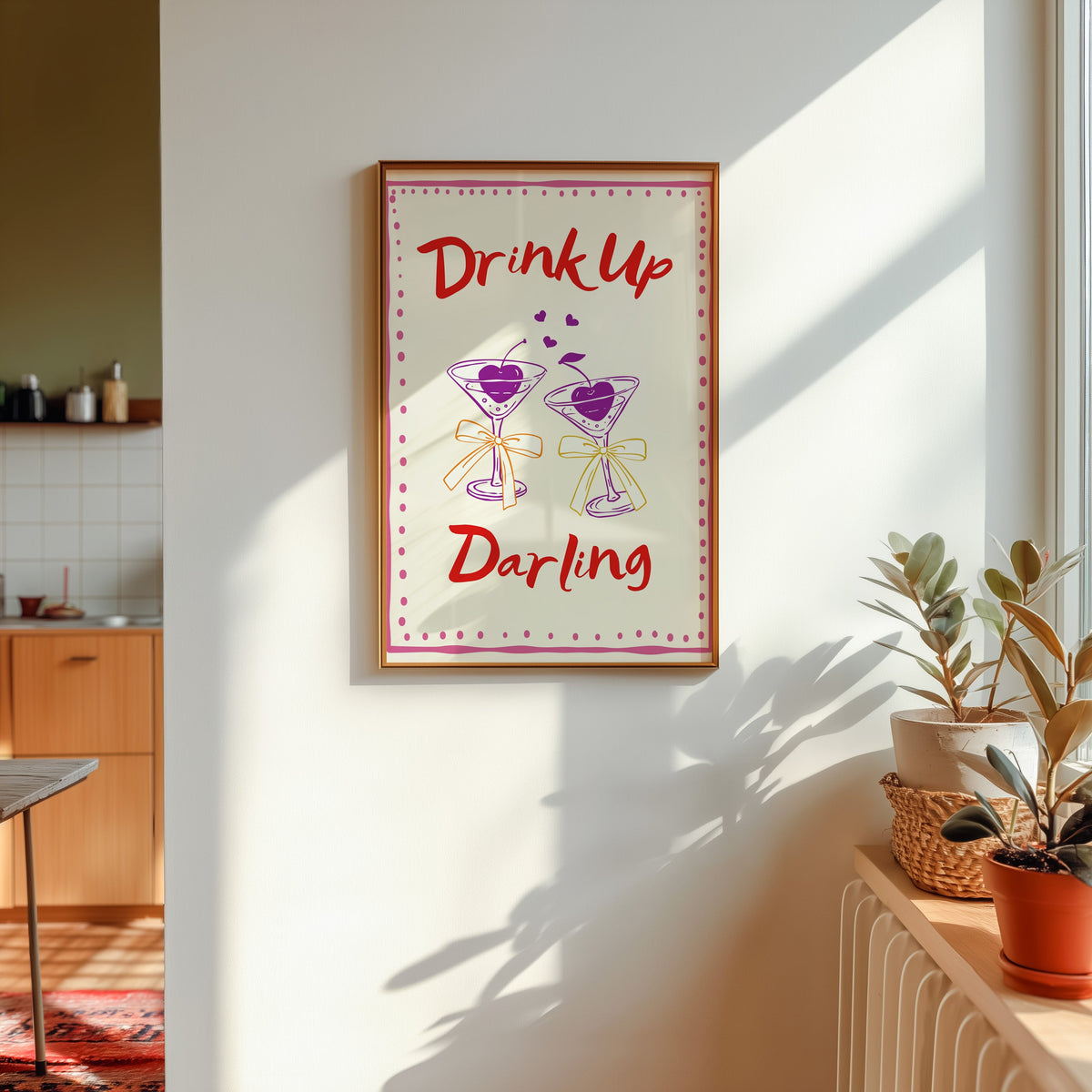Drink Up Darling print in a gold frame in sunlight with plants on the ledge and a kitchen beyond