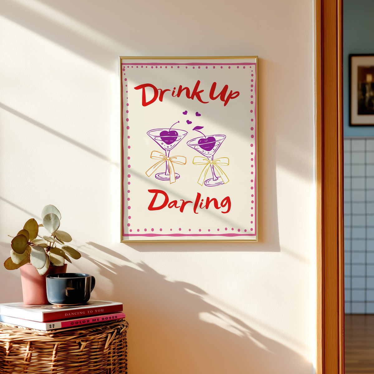 Drink Up Darling print in a gold frame above a basket of books and a pilea plant