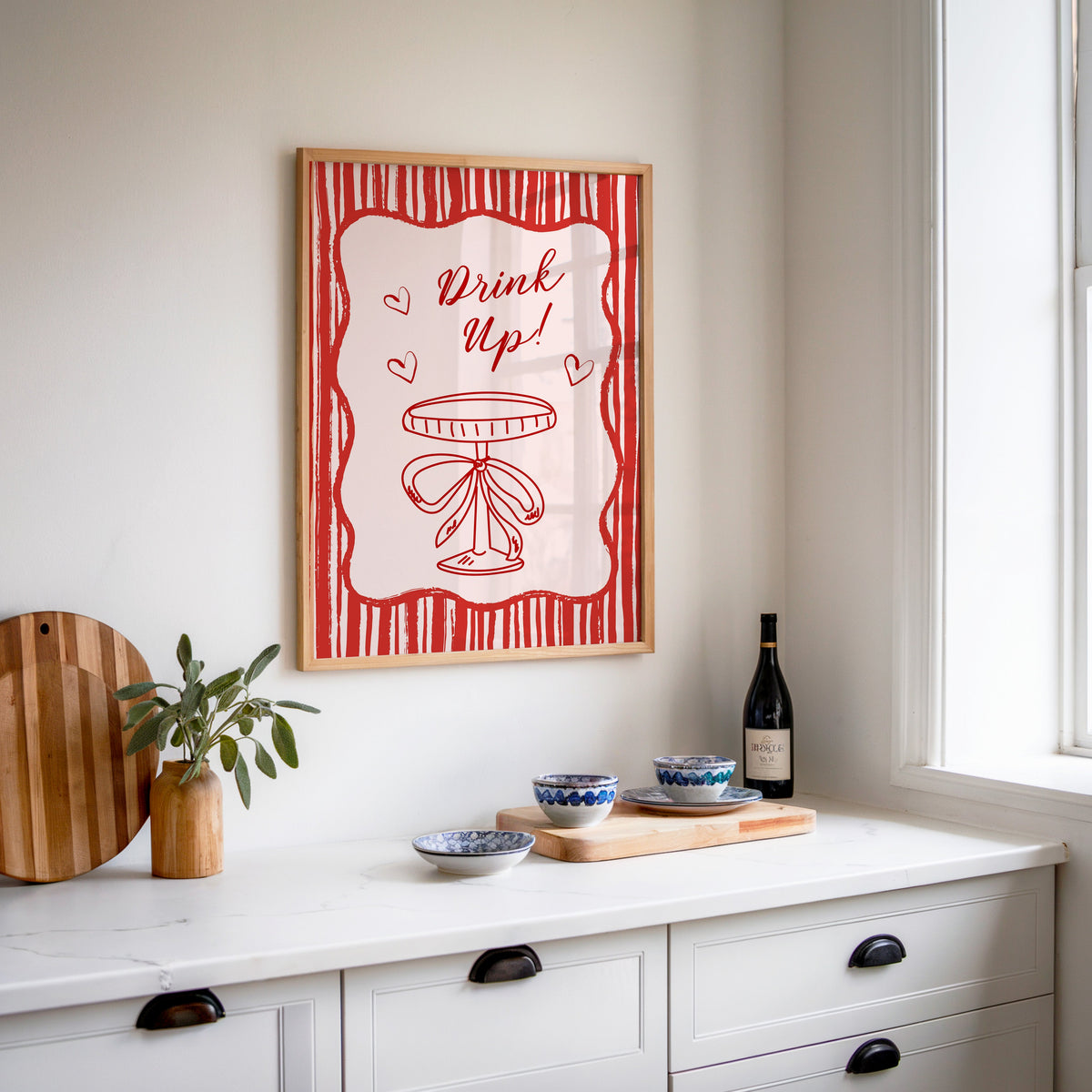 Drink Up coupe print in an oak frame above a white sideboard with a bottle of wine