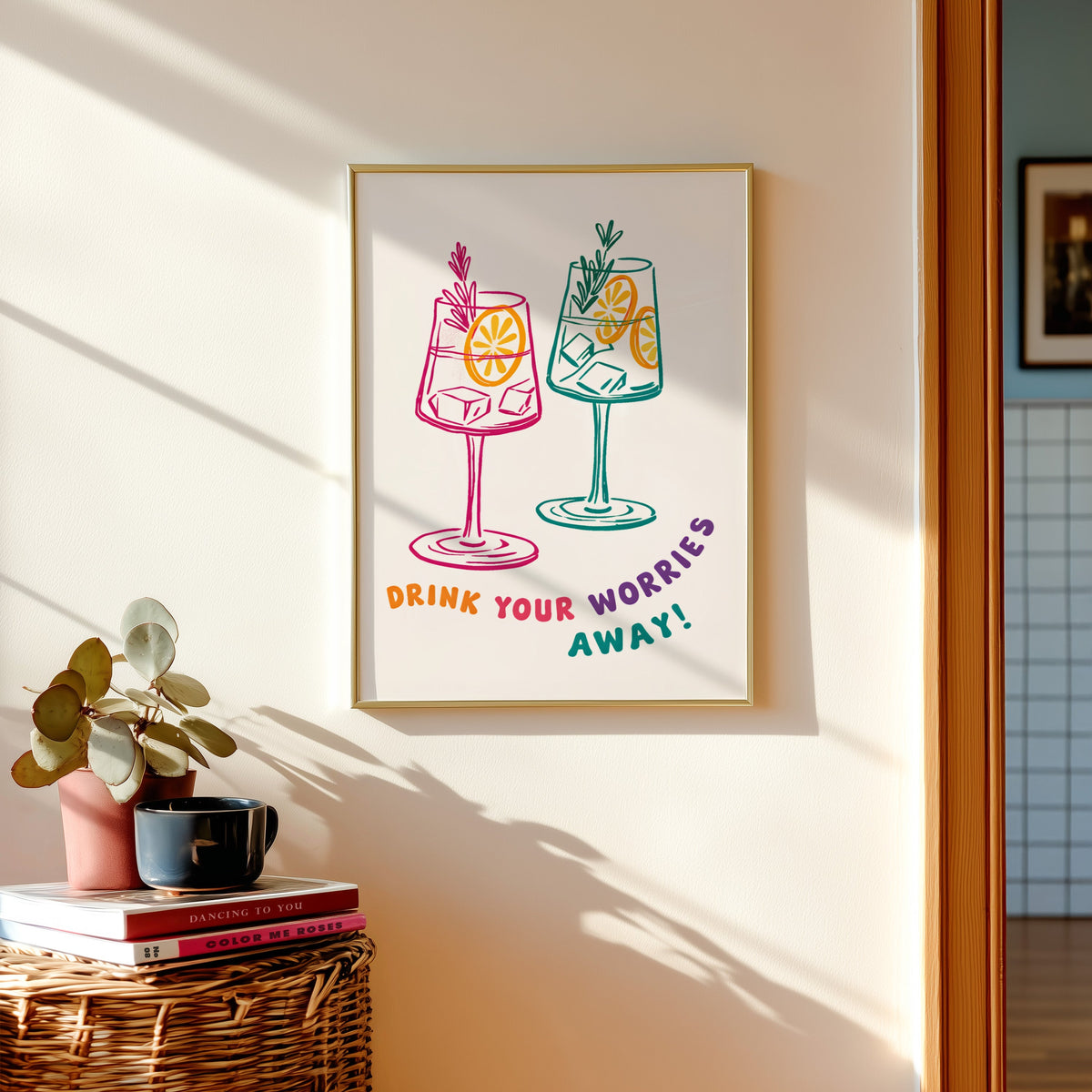 Drink Your Worries Away print shown in a gold frame above a basket of books and a pilea plant
