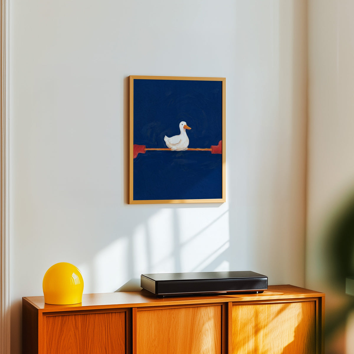 Duck with long legs print in a light oak frame above a teak sideboard with a yellow lamp
