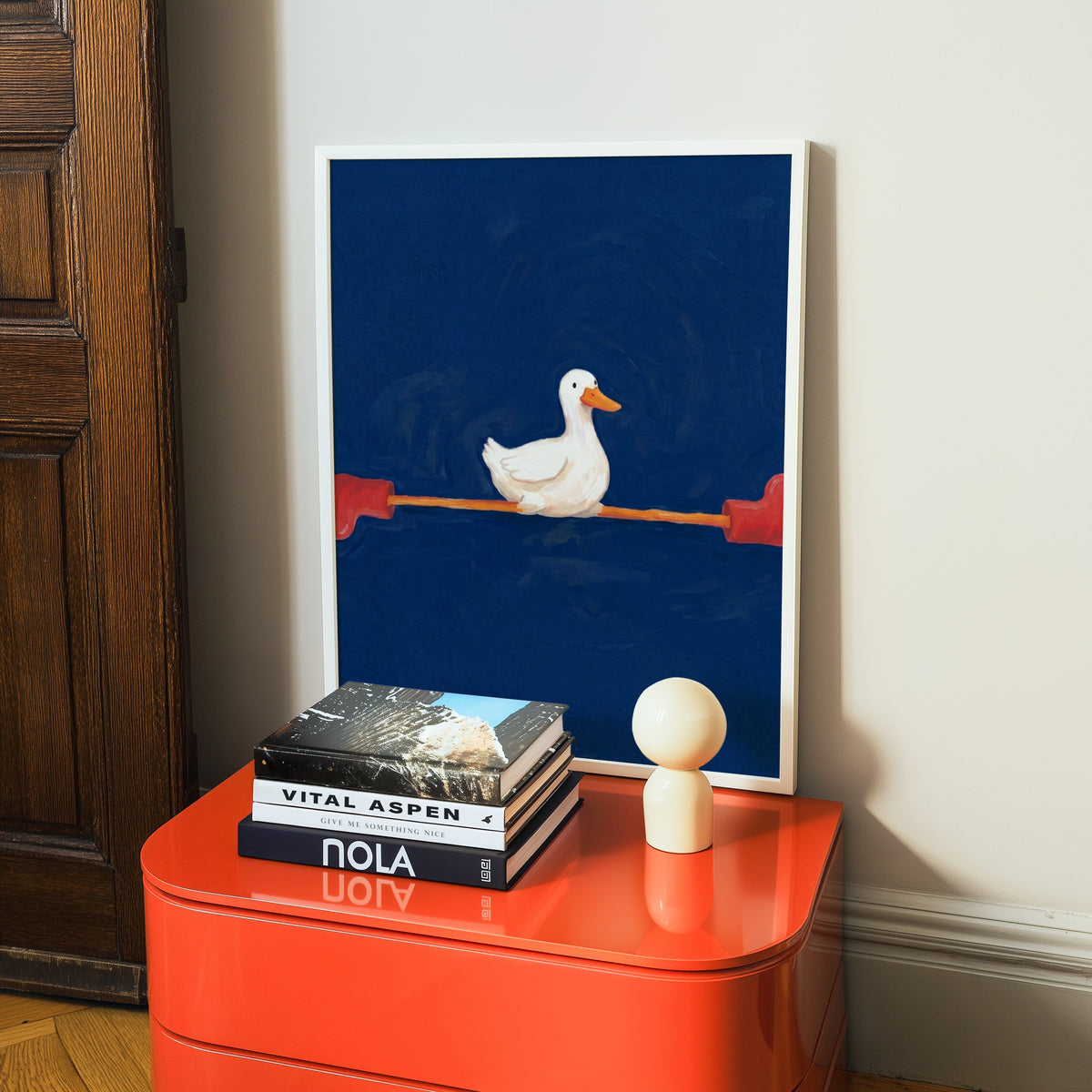 Funny duck print in a white frame on a glossy orange cabinet with books