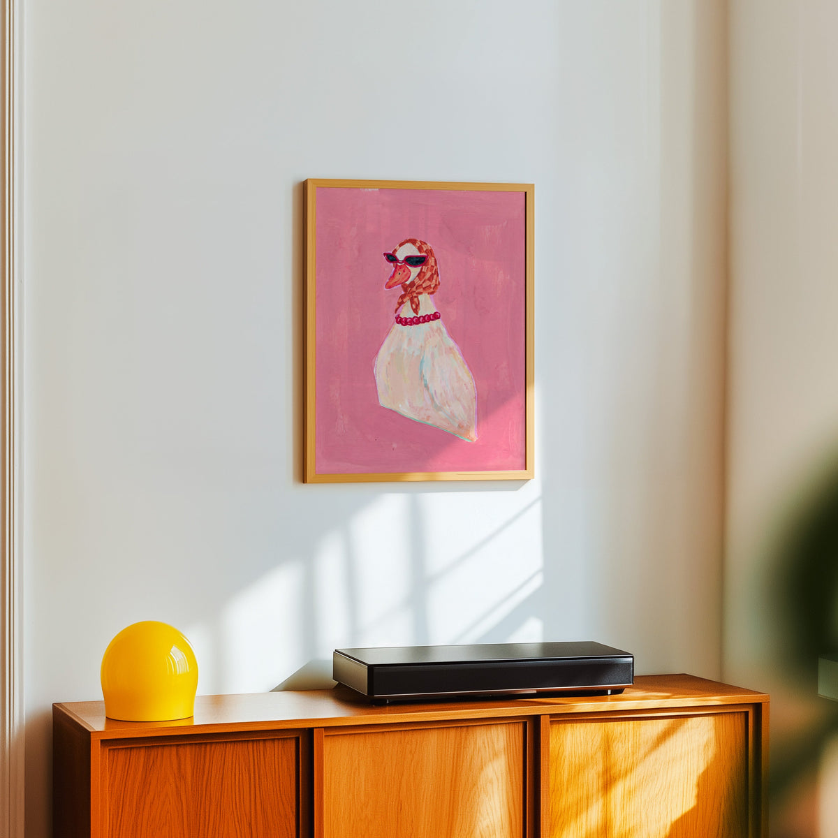 Pink duck fashion print in a light oak frame above a teak sideboard with a yellow lamp