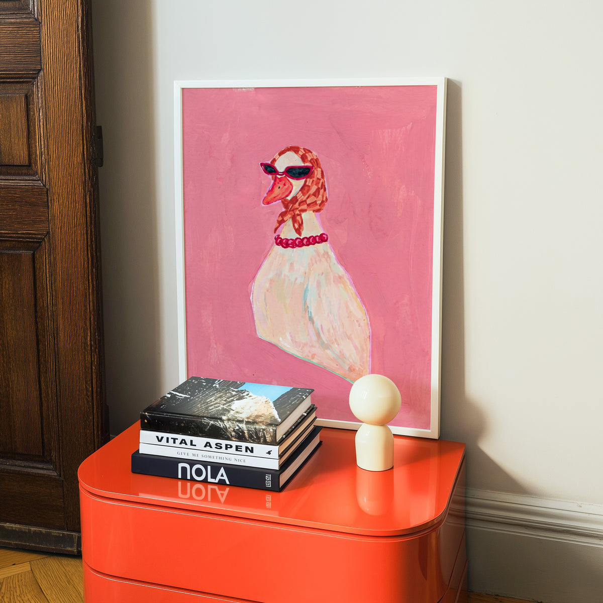 Funny duck print in a white frame on a glossy orange cabinet with books