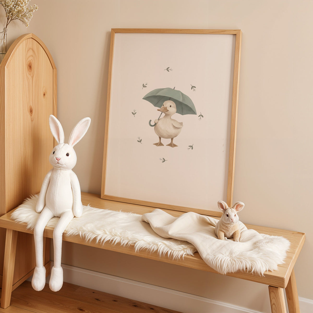 Cream duck holding a sage umbrella nursery print in an oak frame on a sheepskin bench beside a white plush bunny