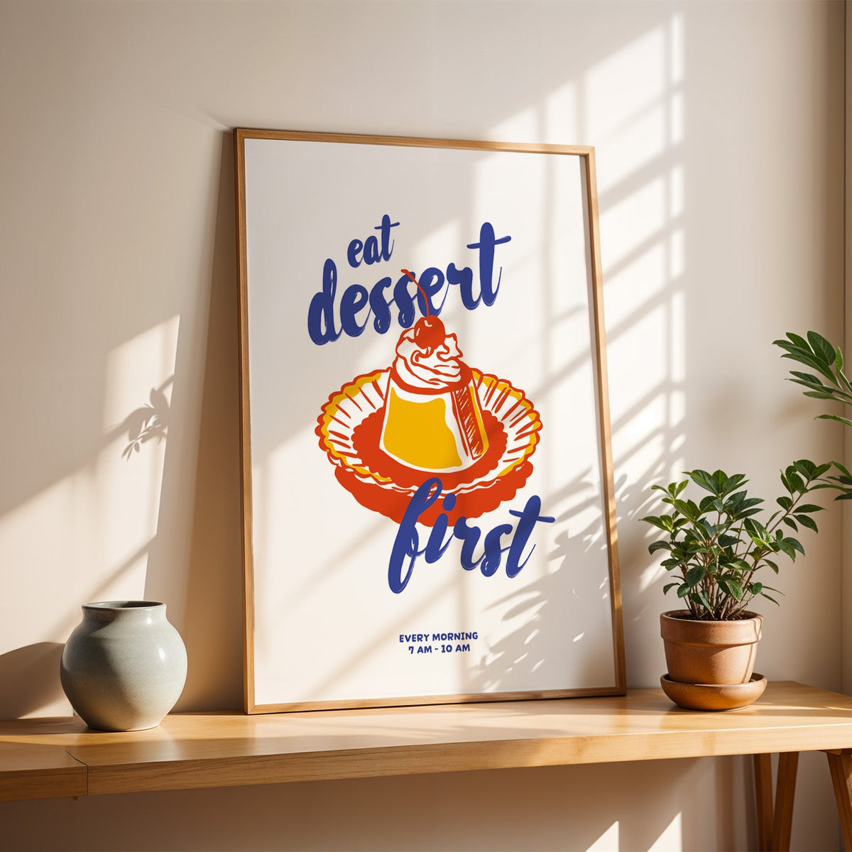 Eat Dessert First poster in a slim oak frame on a sunlit wood console with a stoneware vase