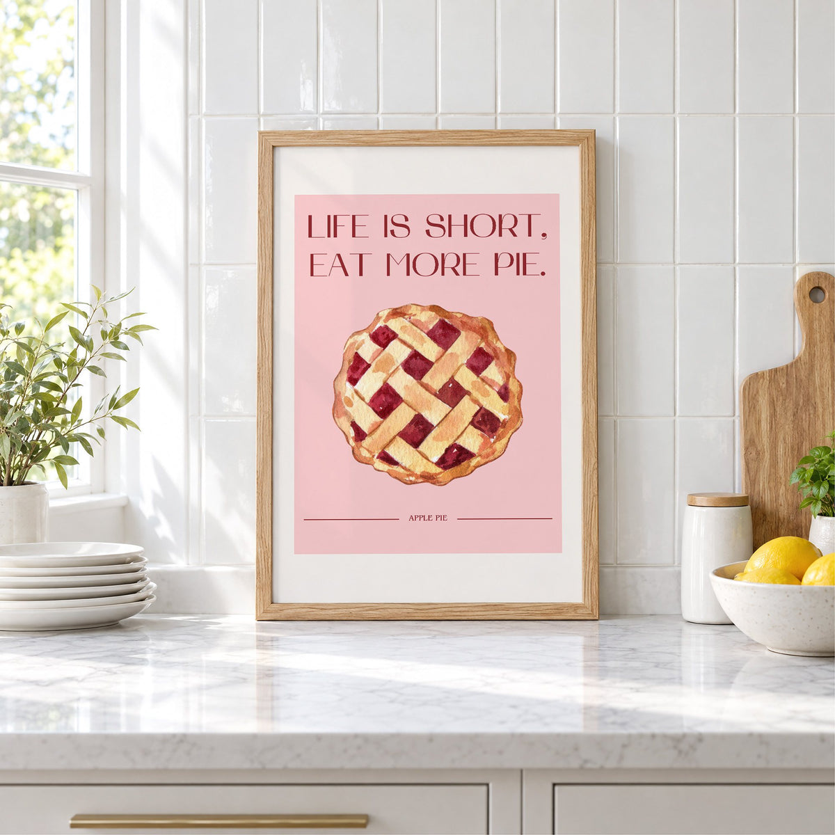 Eat More Pie watercolor print in an oak frame leaning on a marble counter by white tiles and lemons