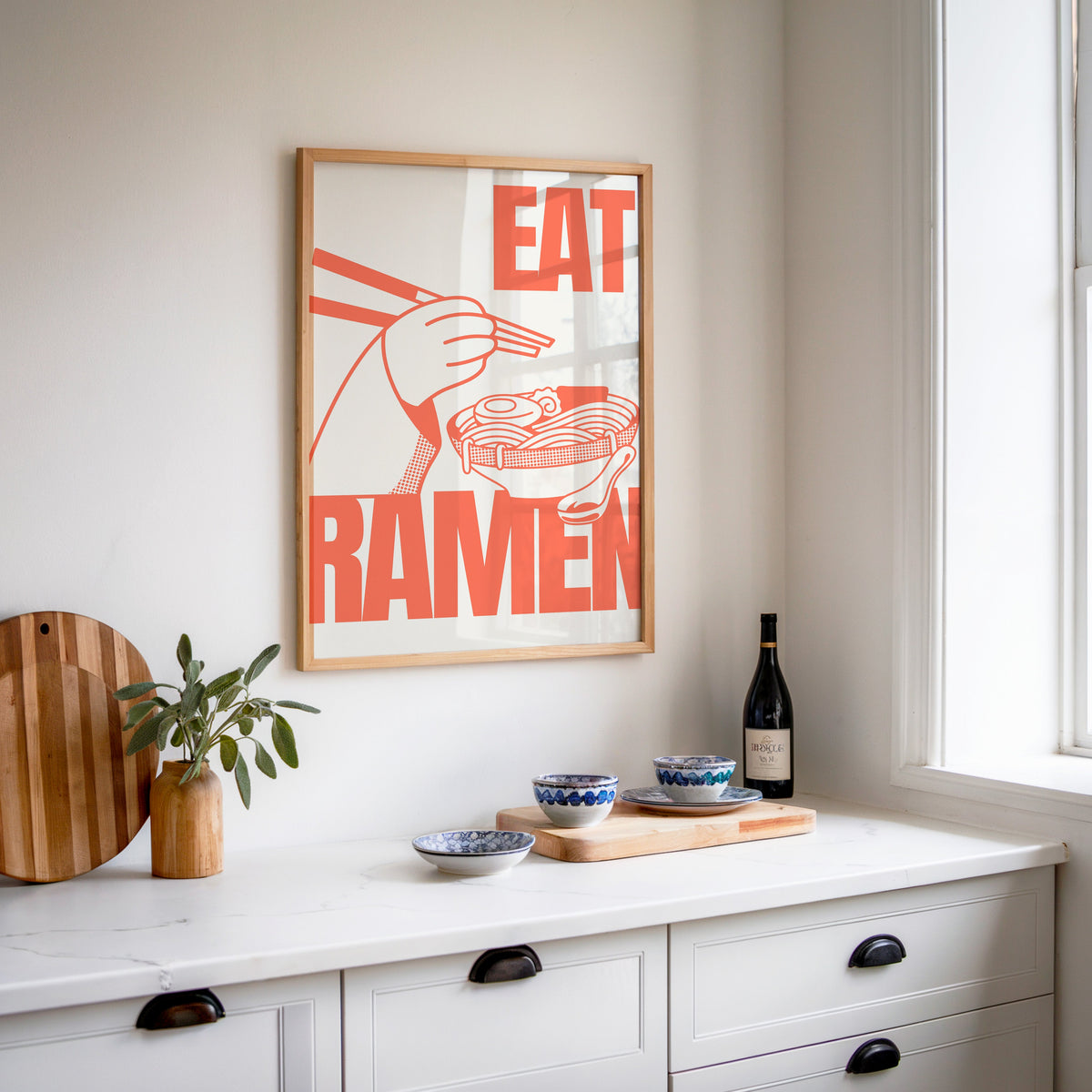 Eat Ramen print in an oak frame above a white sideboard with a bottle of wine