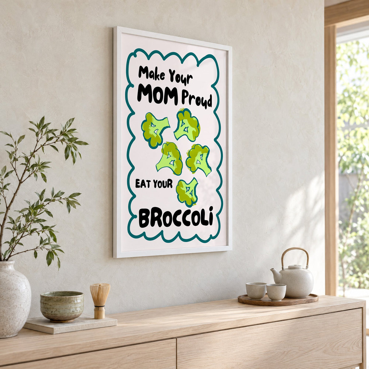 Eat Your Broccoli pink kitchen print in a white frame above a pale wood console with a matcha tea set