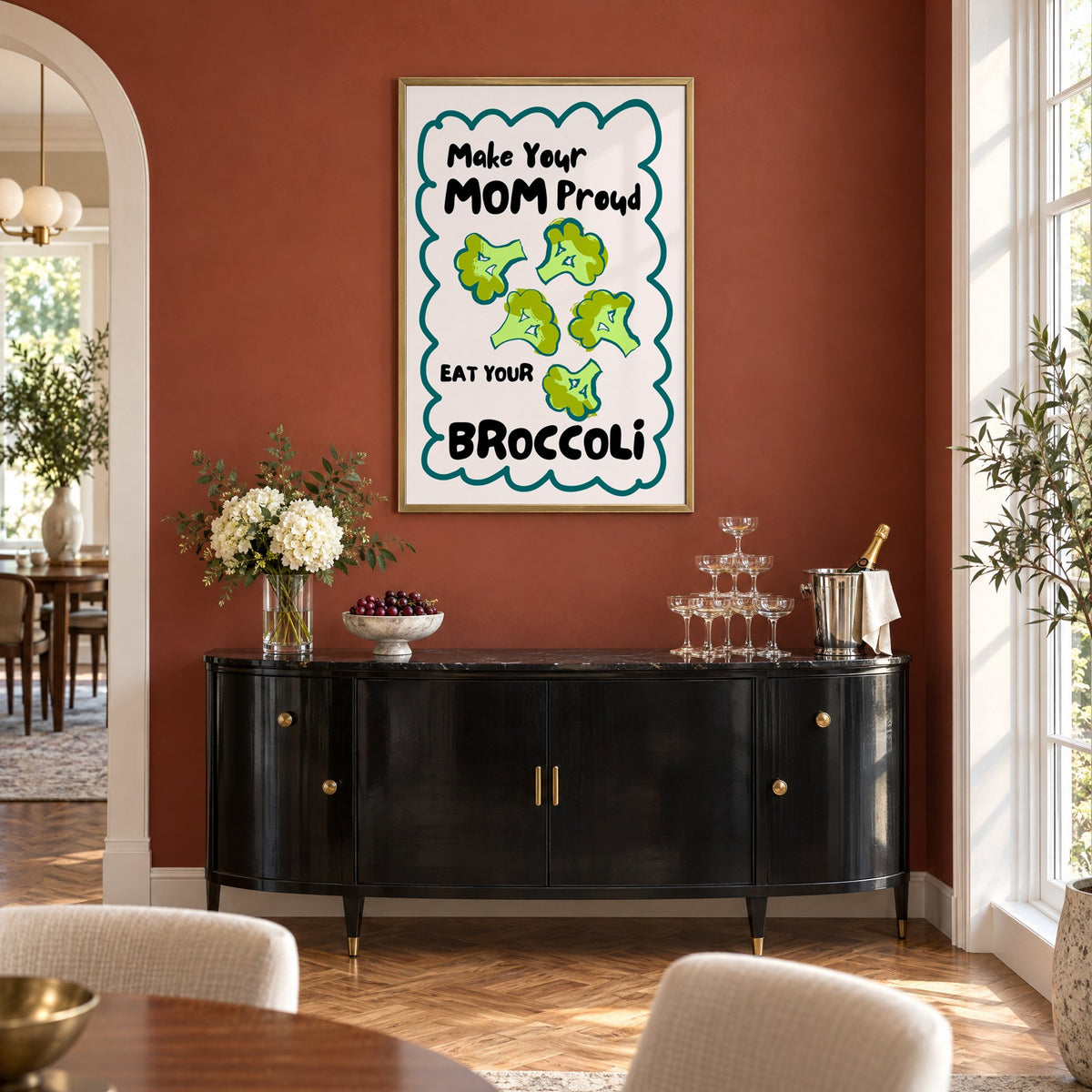 Eat Your Broccoli pink kitchen print in a brass frame on a terracotta wall above a black buffet with a champagne tower