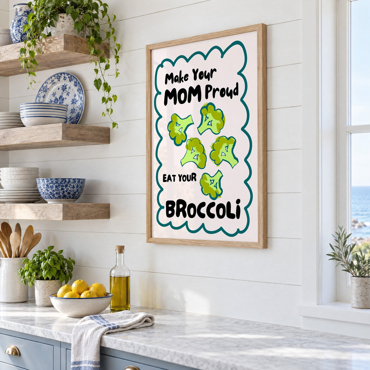 Eat Your Broccoli pink kitchen print in an oak frame in a white coastal kitchen beside blue and white plates