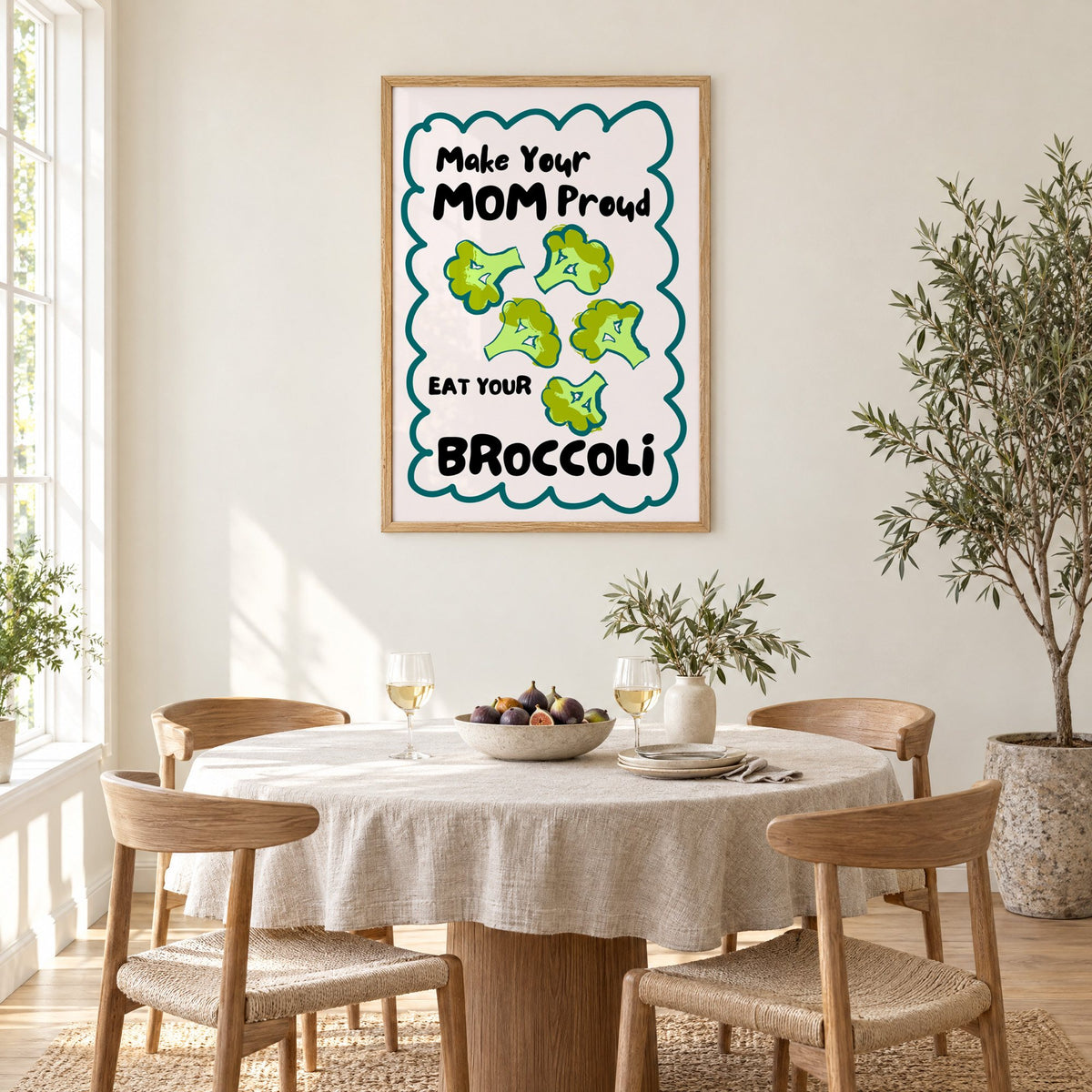 Eat Your Broccoli pink kitchen print in an oak frame above a round linen-covered dining table with figs and wine