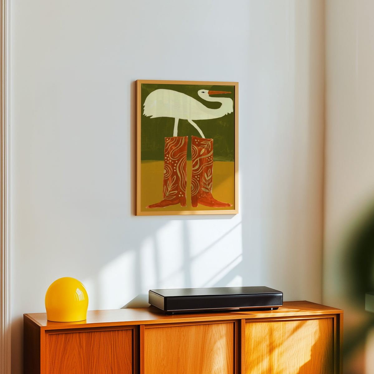 Bird wearing cowboy boots print in a light oak frame above a teak sideboard with a yellow lamp