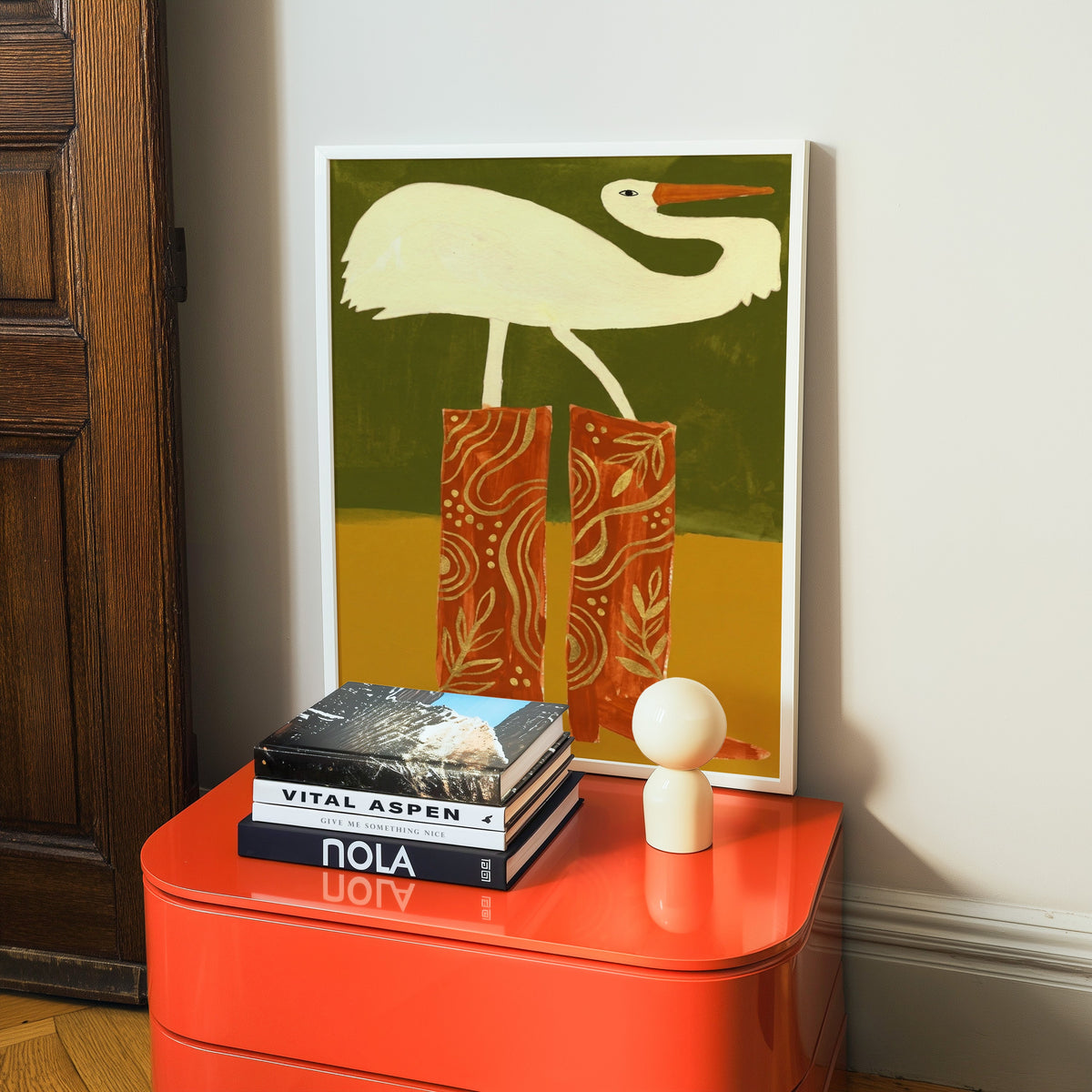 Quirky bird in red boots print in a white frame on a glossy orange cabinet with books