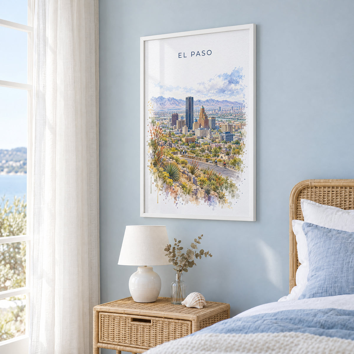 El Paso watercolor print in a white frame on a pale blue wall beside a rattan bed in a coastal bedroom