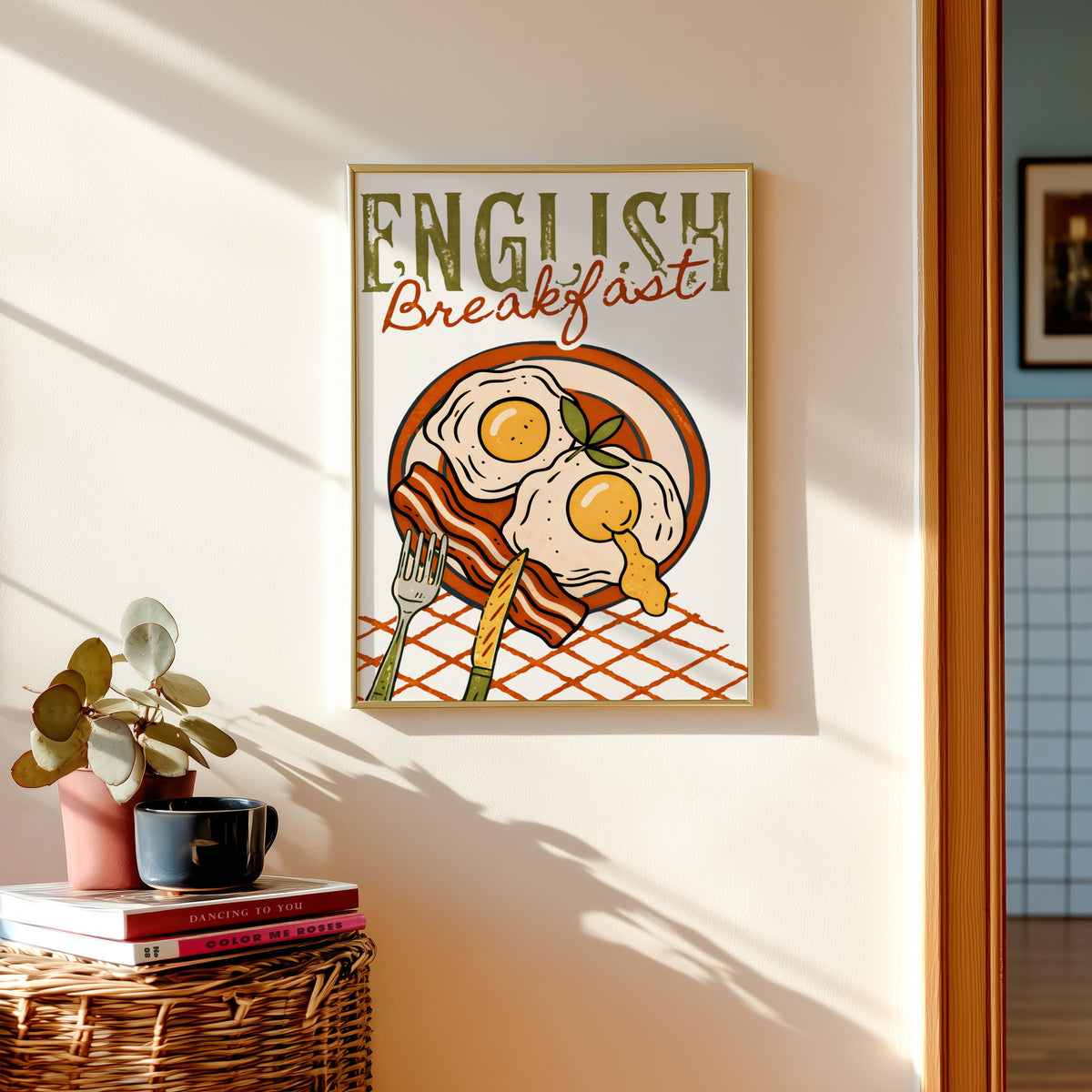 English breakfast print in a gold frame above a basket of books and a pilea plant
