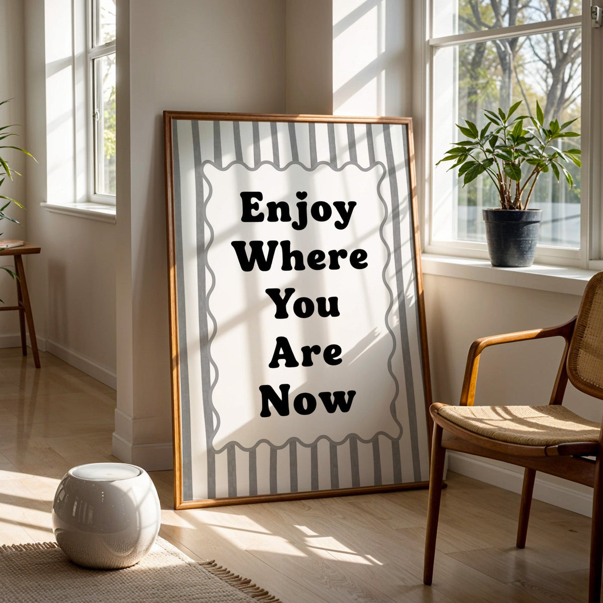 Enjoy where you are now striped quote print in a walnut frame on a wood floor beside a cane chair and sunny window