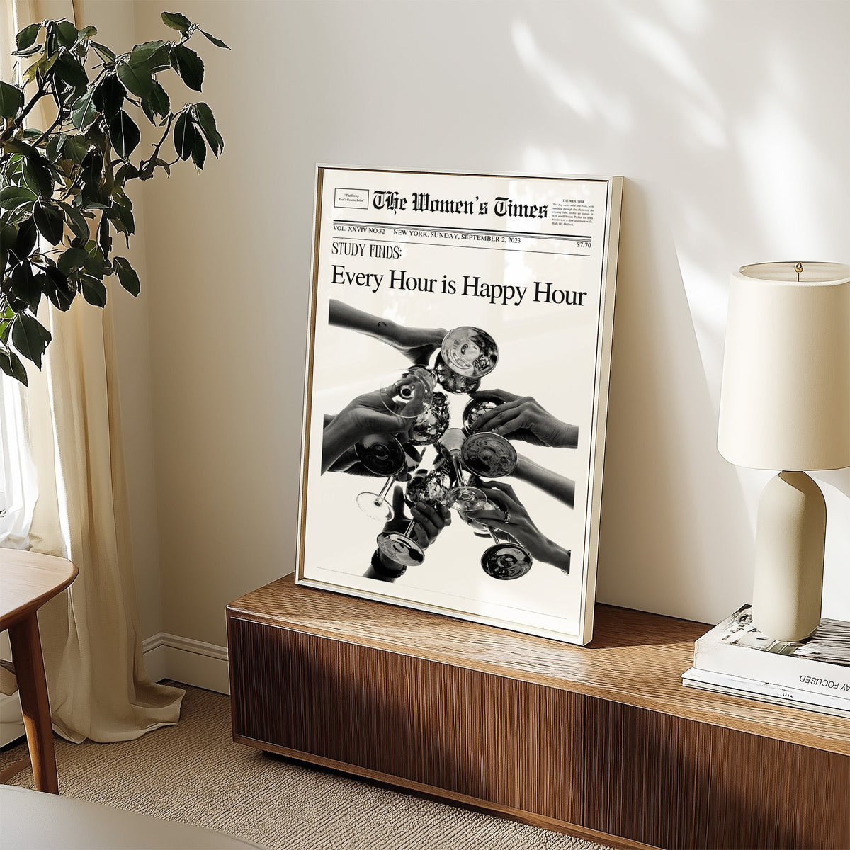 Mock front page with hands toasting cocktail glasses print in a white frame on a walnut sideboard beside a ceramic lamp