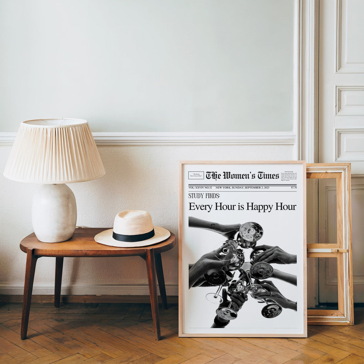Mock front page with hands toasting cocktail glasses print in a pine frame on a wood floor beside a lamp and straw hat