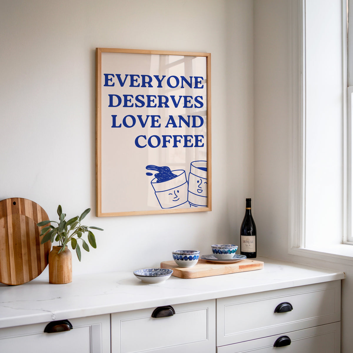 Everyone Deserves Love and Coffee print in an oak frame above a white sideboard with a bottle of wine