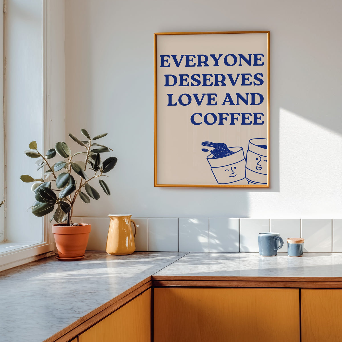 Everyone Deserves Love and Coffee print shown in a gold frame above a marble counter in a tan kitchen