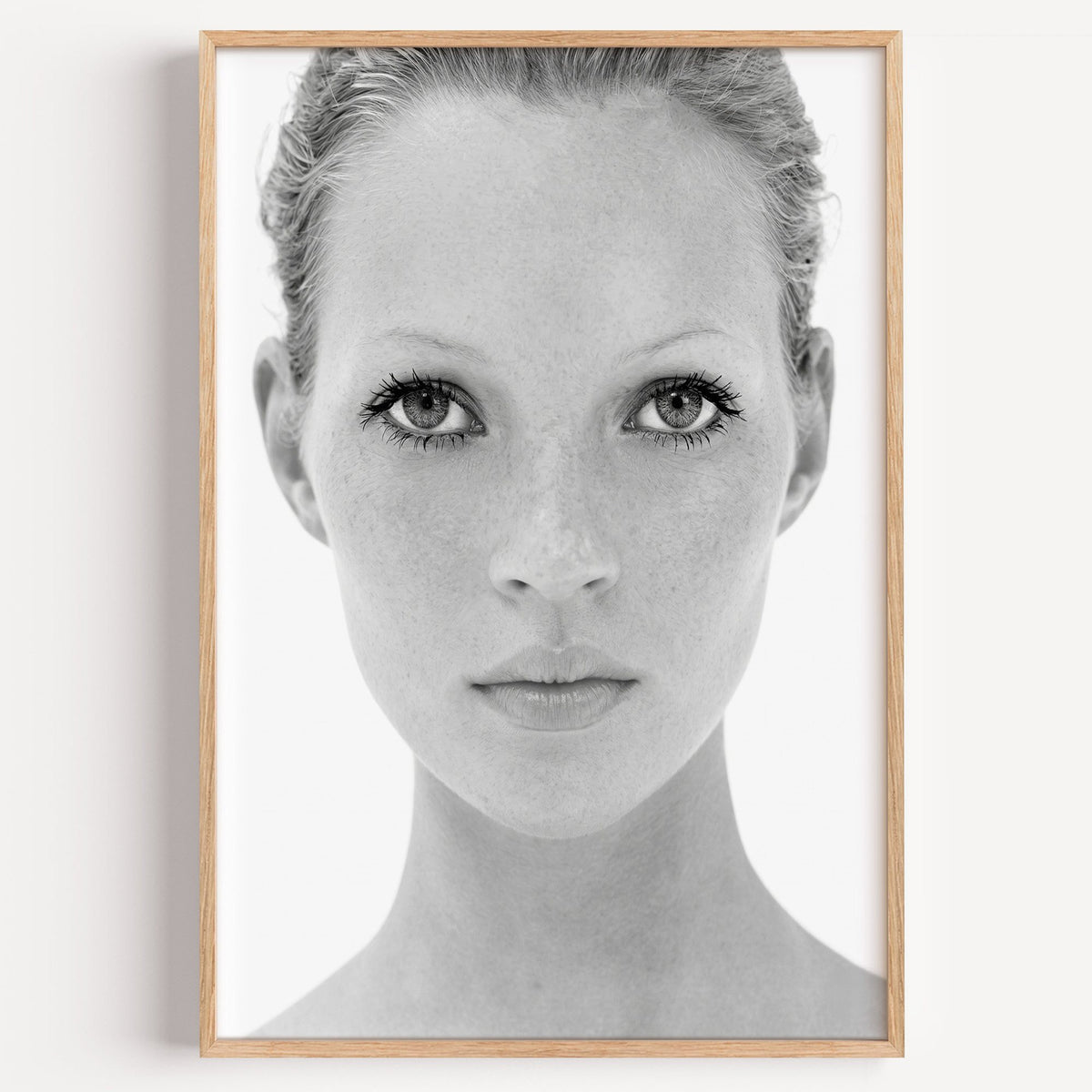 Bright face closeup with dark lashes print in a light oak frame on a plain white wall