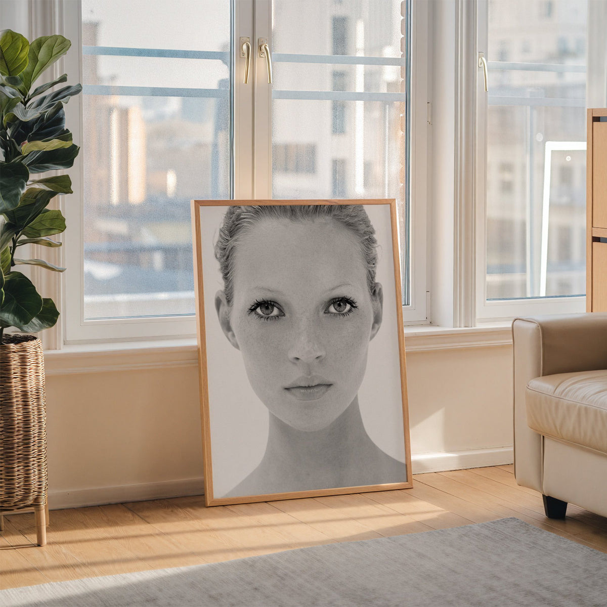 Bright face closeup with dark lashes print in an oak frame leaning on the floor beside a fiddle leaf fig