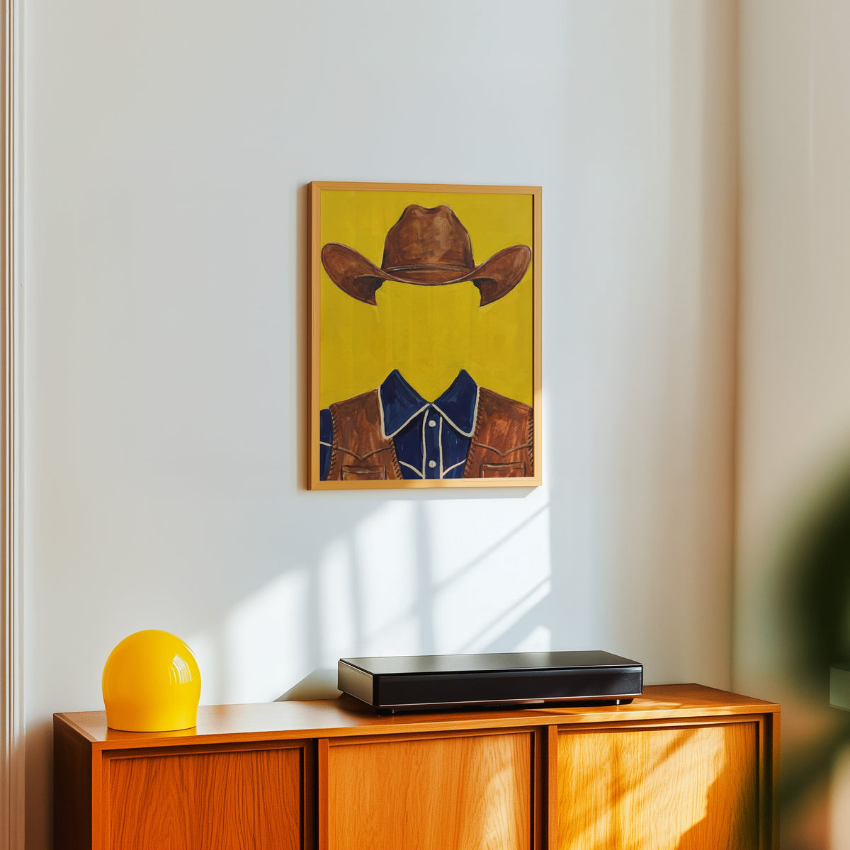 Yellow cowboy print in a light oak frame above a teak sideboard with a yellow lamp