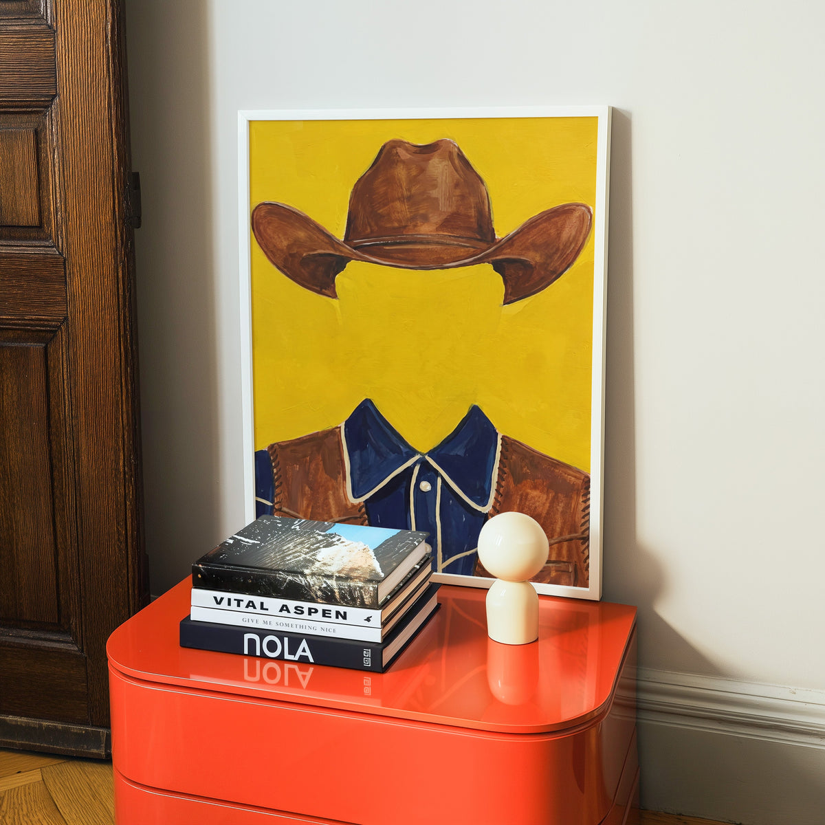 Cowboy hat and vest print in a white frame on a glossy orange cabinet with books