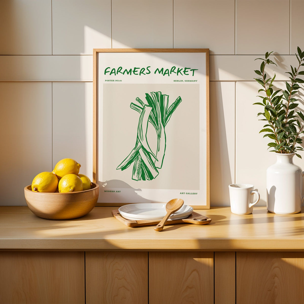 Green leeks print in a slim oak frame leaning on a counter beside a wooden bowl of lemons