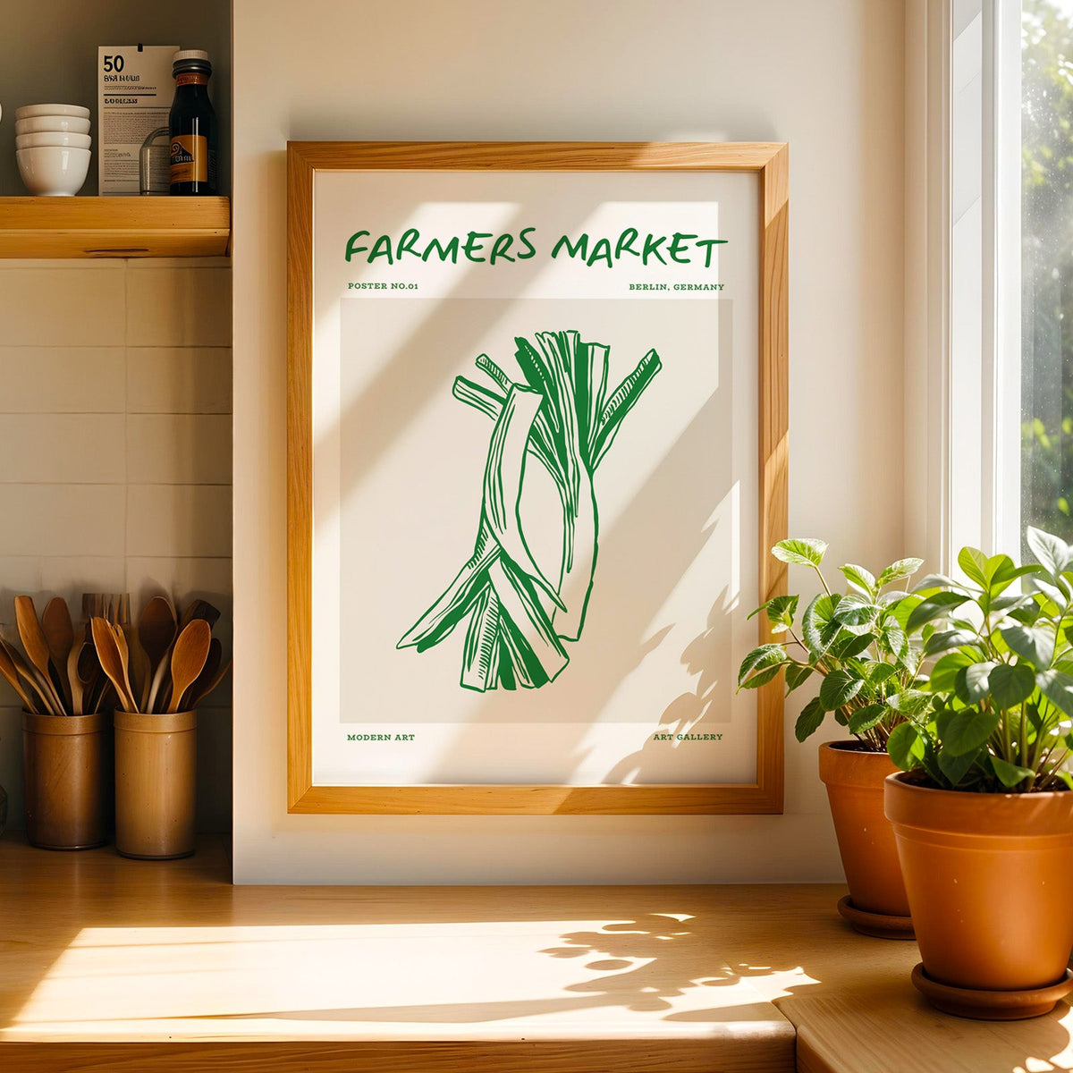 Green leeks print in a light pine frame on a counter next to a ceramic jar of wooden spoons