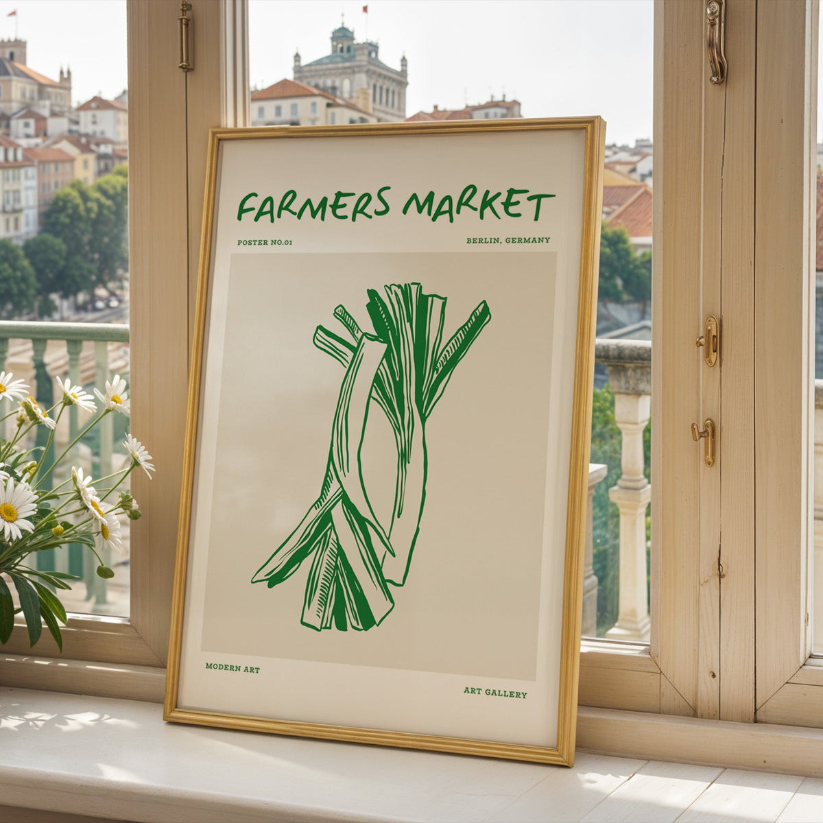Green leeks print in a gold frame on a window sill above a European hillside town
