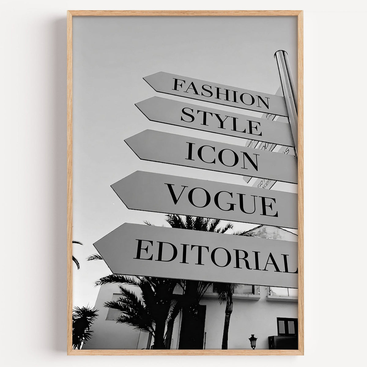 Street signpost with fashion words above palm trees print in a light oak frame on a plain white wall