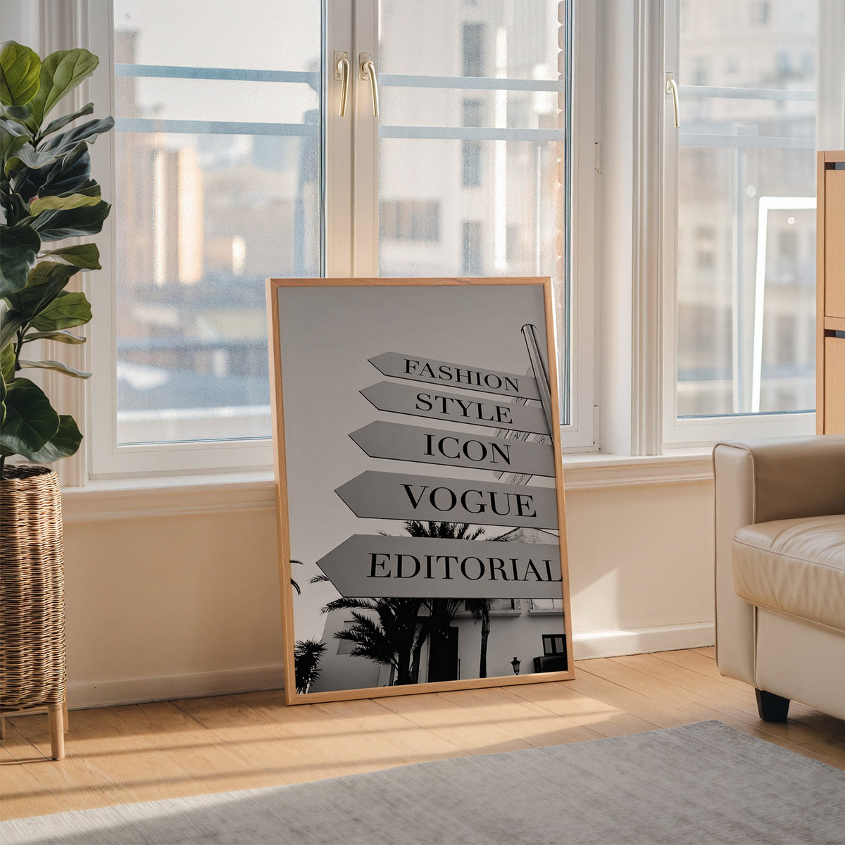 Street signpost with fashion words above palm trees print in an oak frame leaning on the floor beside a fiddle leaf fig