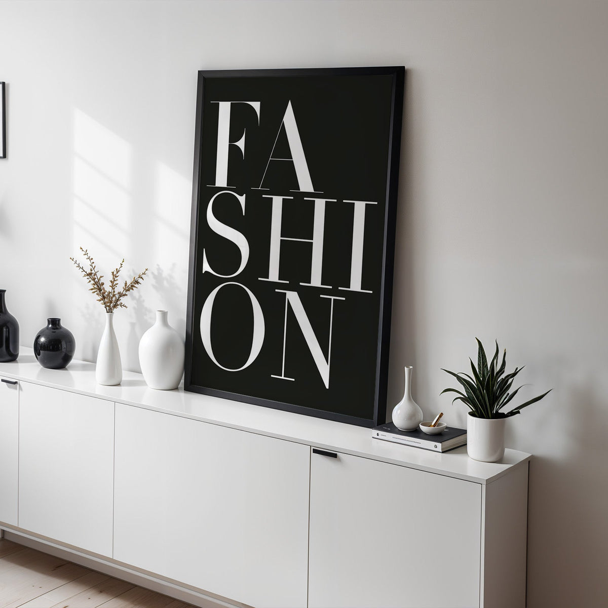 Stacked serif FASHION lettering on charcoal print in a black frame on a white sideboard with black and white vases