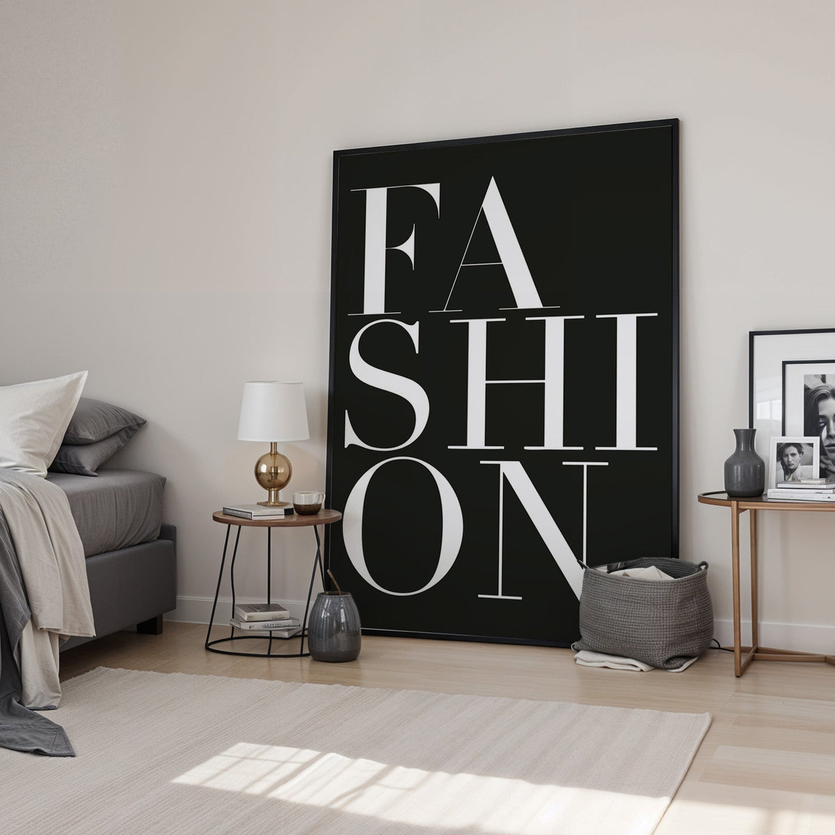 Stacked serif FASHION lettering on charcoal print in a black frame leaning in a bedroom beside a gold lamp