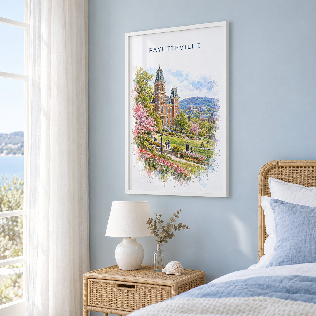 Fayetteville, Arkansas watercolor print in a white frame on a pale blue wall beside a rattan bed in a coastal bedroom