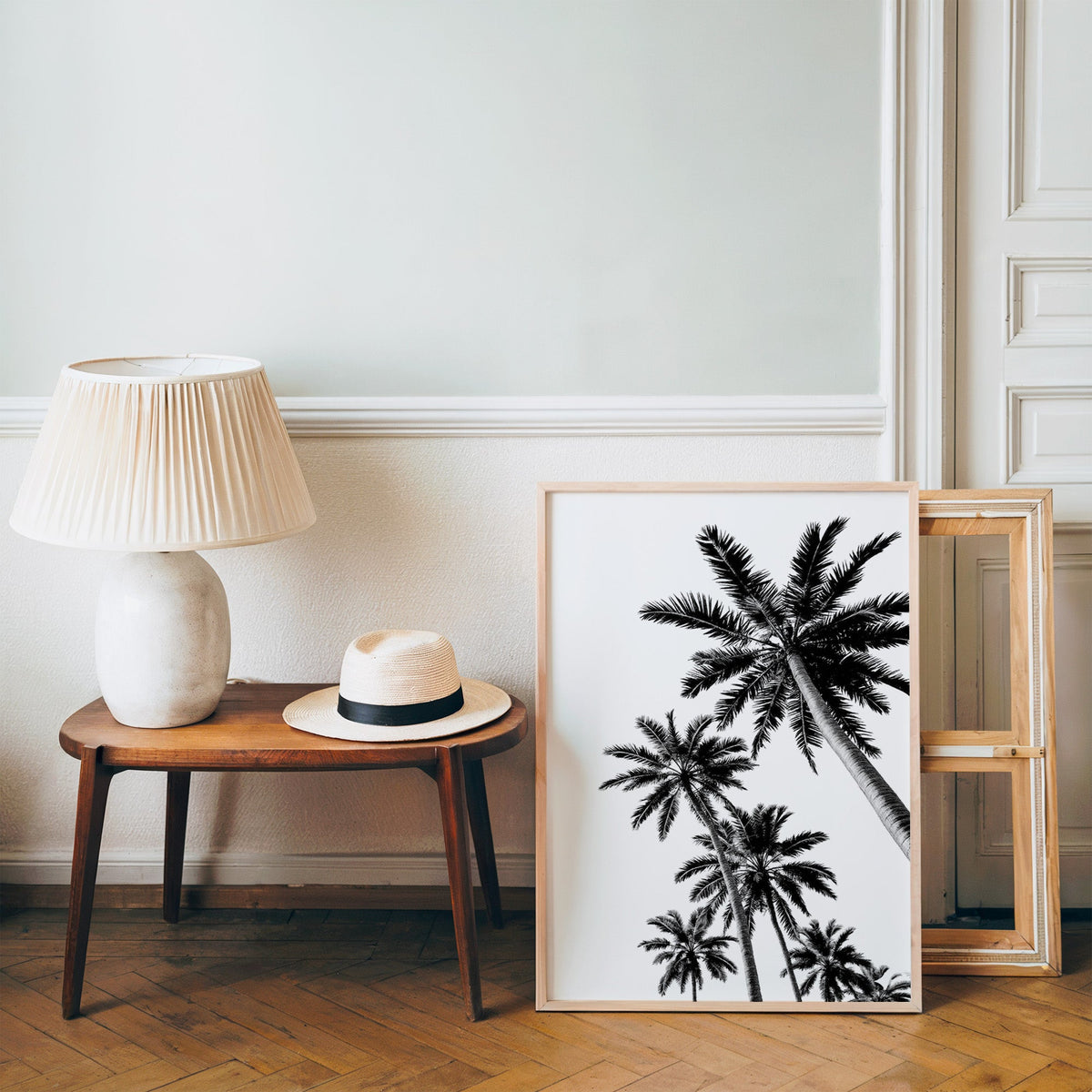 Feather leaf palms on a white sky print in a pine frame on a wood floor beside a lamp and straw hat