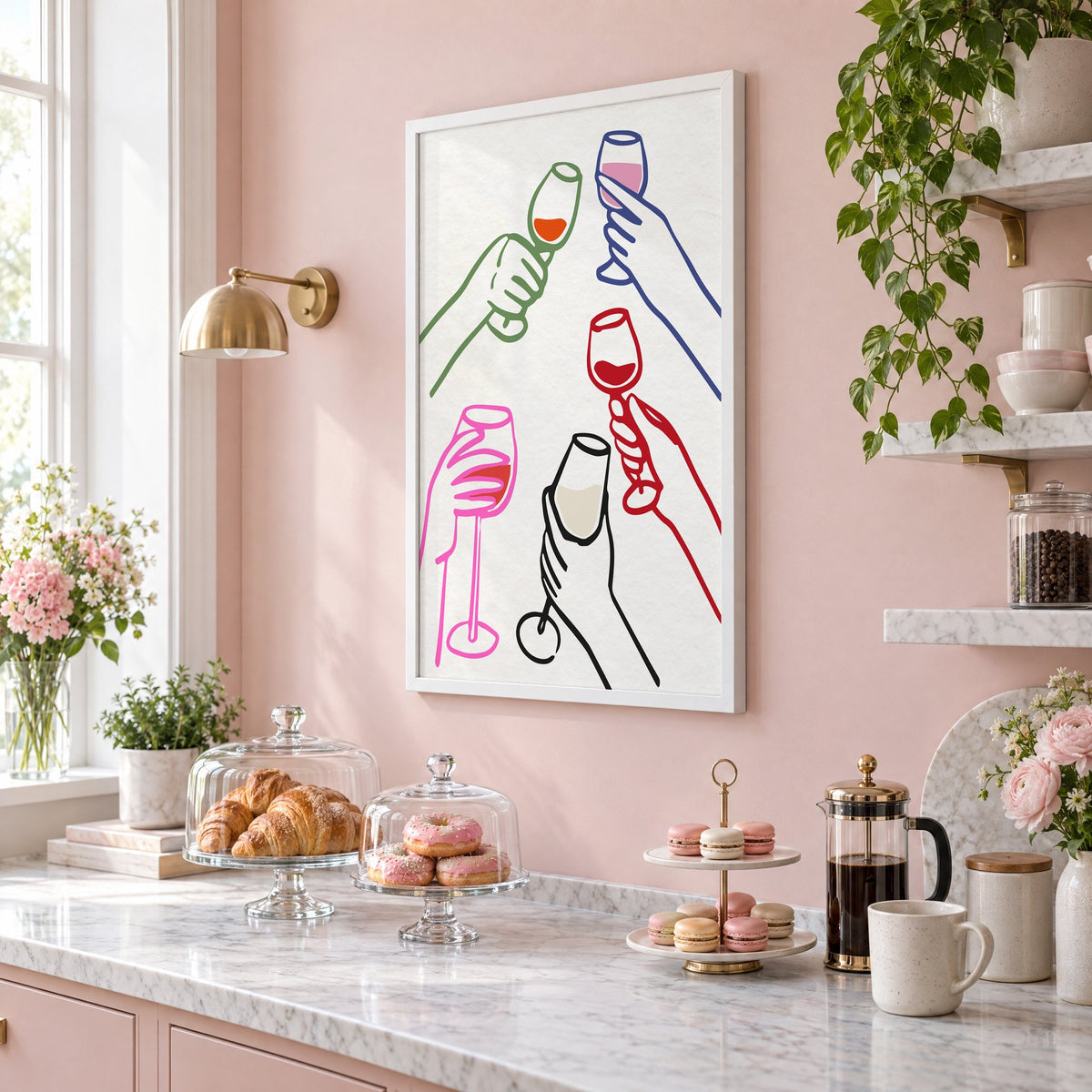 Five hands multicolor wine toast print in a white frame on a blush pink wall above a patisserie counter with croissants and macarons, seen at an angle