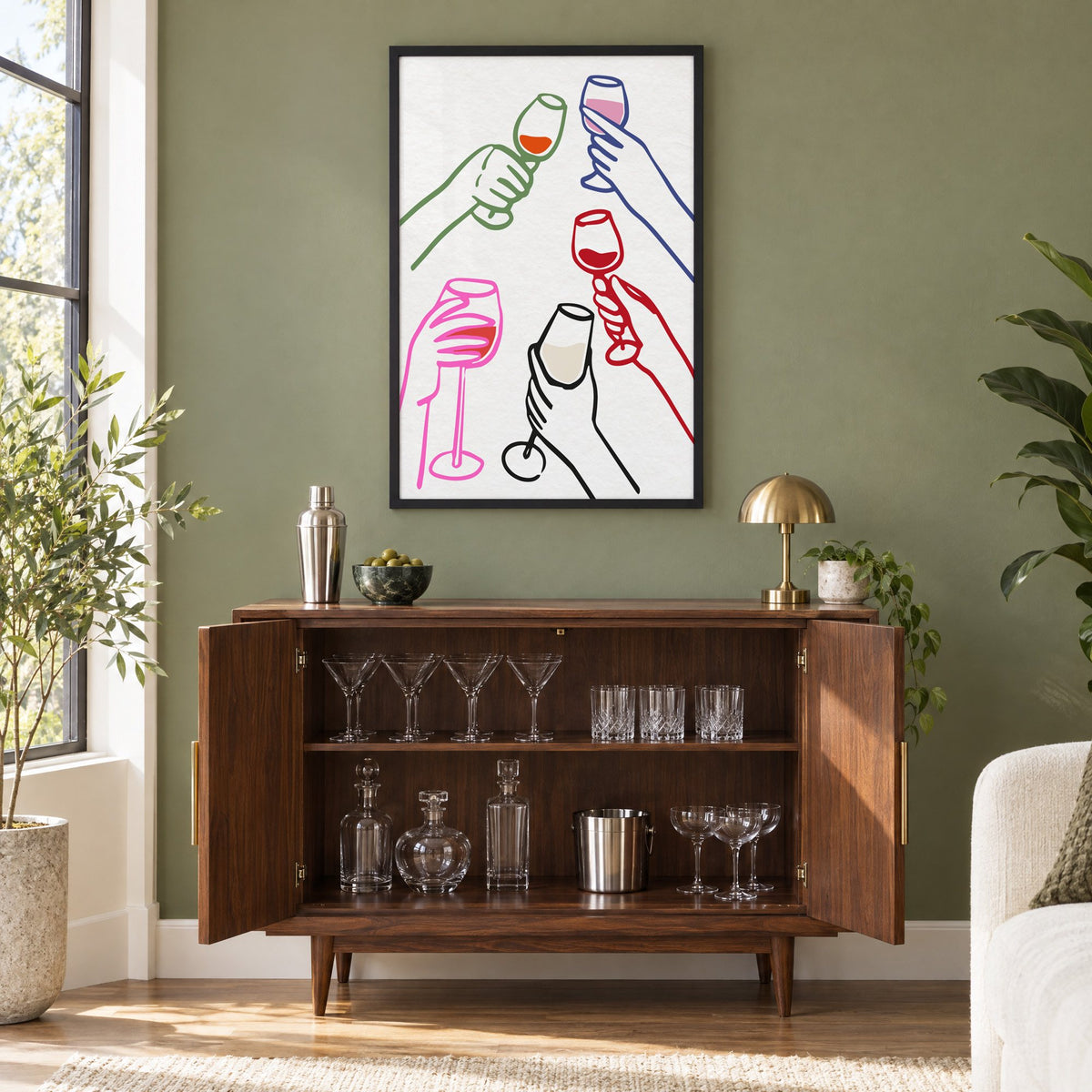 Five hands multicolor wine toast print in a black frame on an olive green wall above a walnut bar cabinet with glassware
