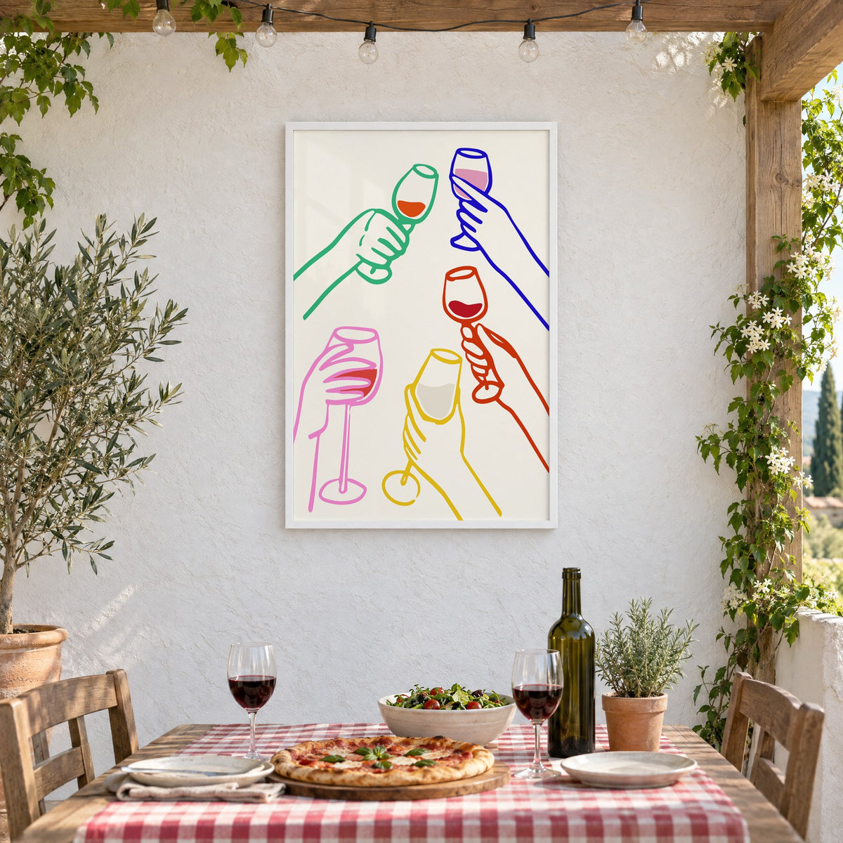 Five hands colorful line wine toast print in a white frame on a stucco wall above a checked-cloth table with pizza and red wine
