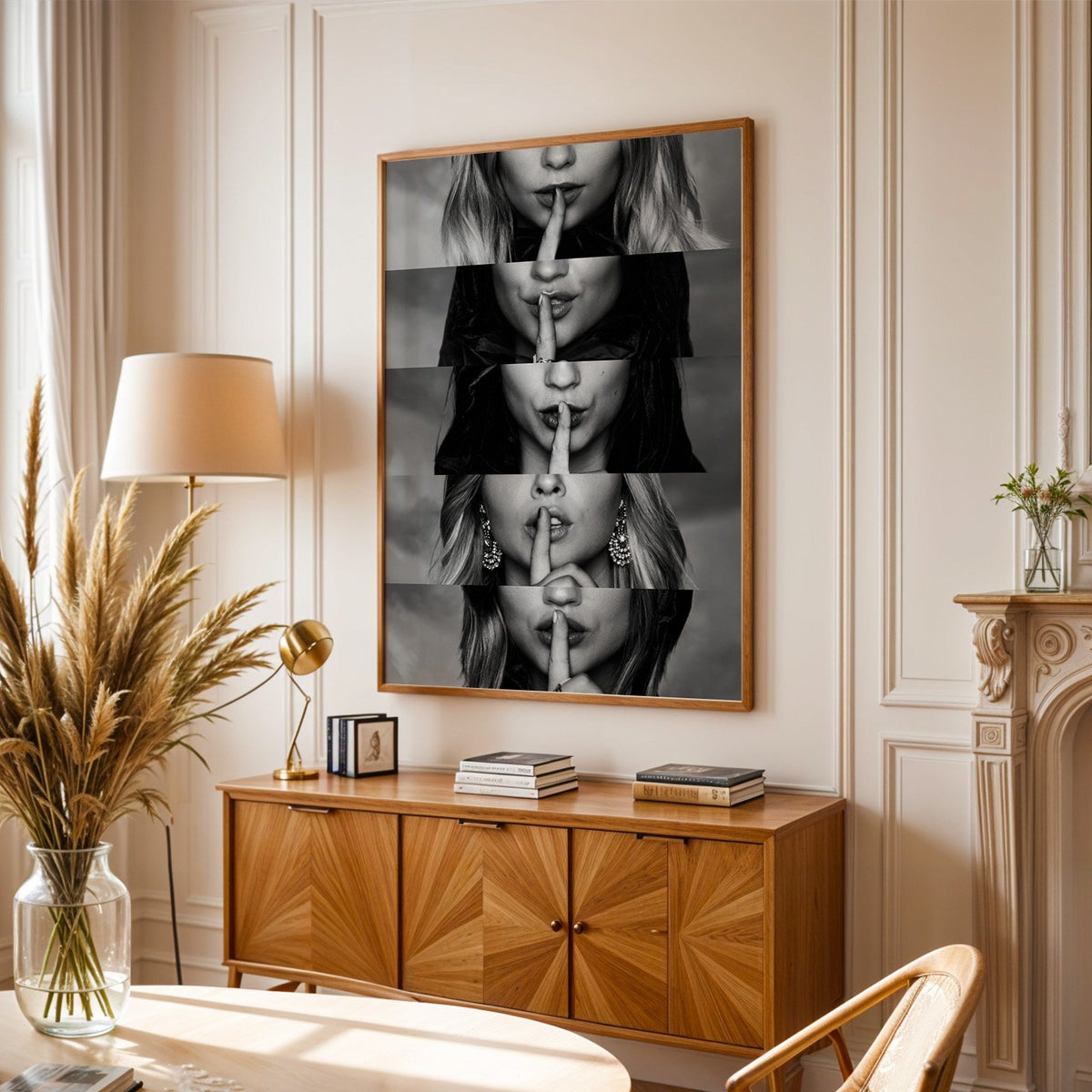 Five stacked strips of finger to lips hush print in an oak frame above a sunburst wood sideboard with pampas grass