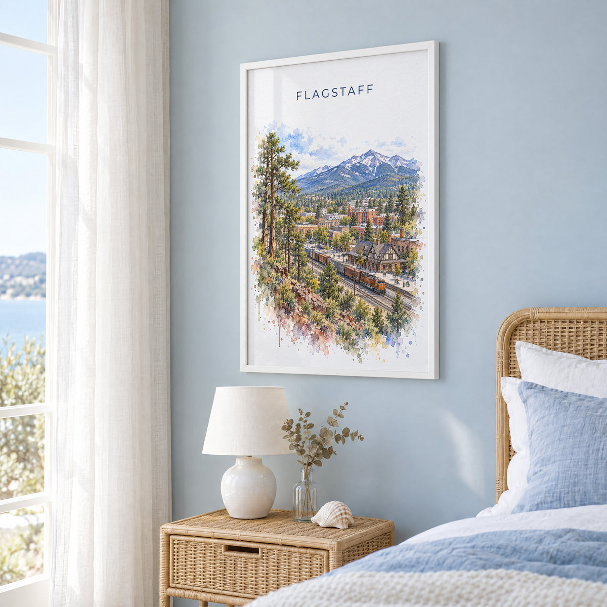 Flagstaff watercolor print in a white frame on a pale blue wall beside a rattan bed in a coastal bedroom