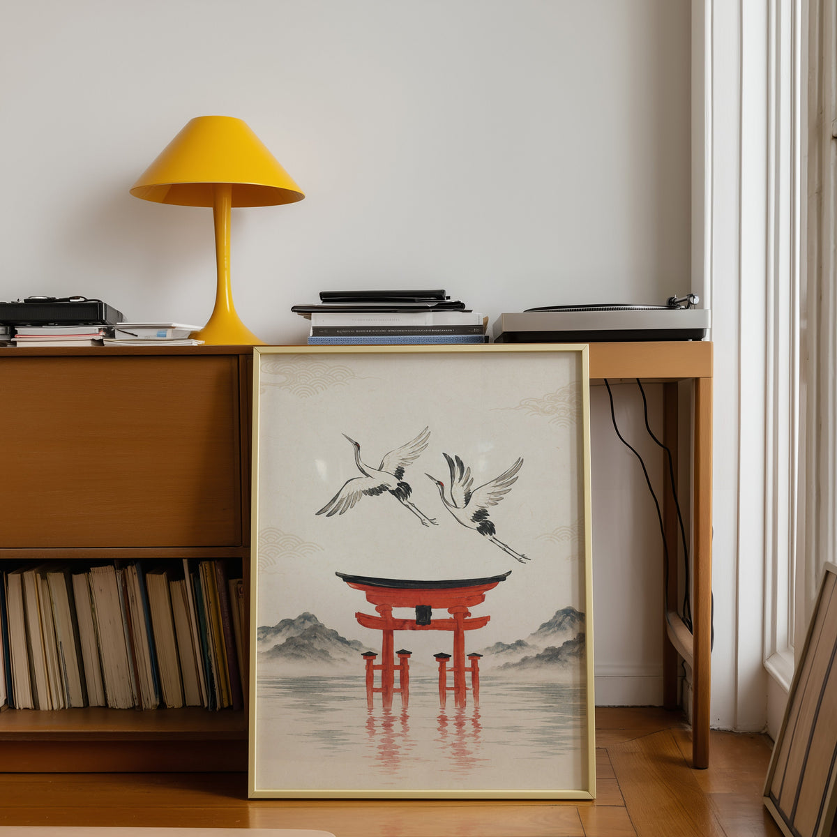 red torii gate with cranes print shown in a gold frame leaning under a teak desk with a yellow lamp
