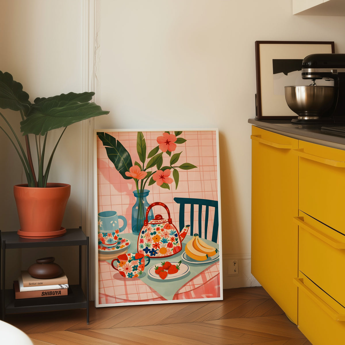 Floral teapot print in a white frame leaning on parquet beside a bright yellow kitchen